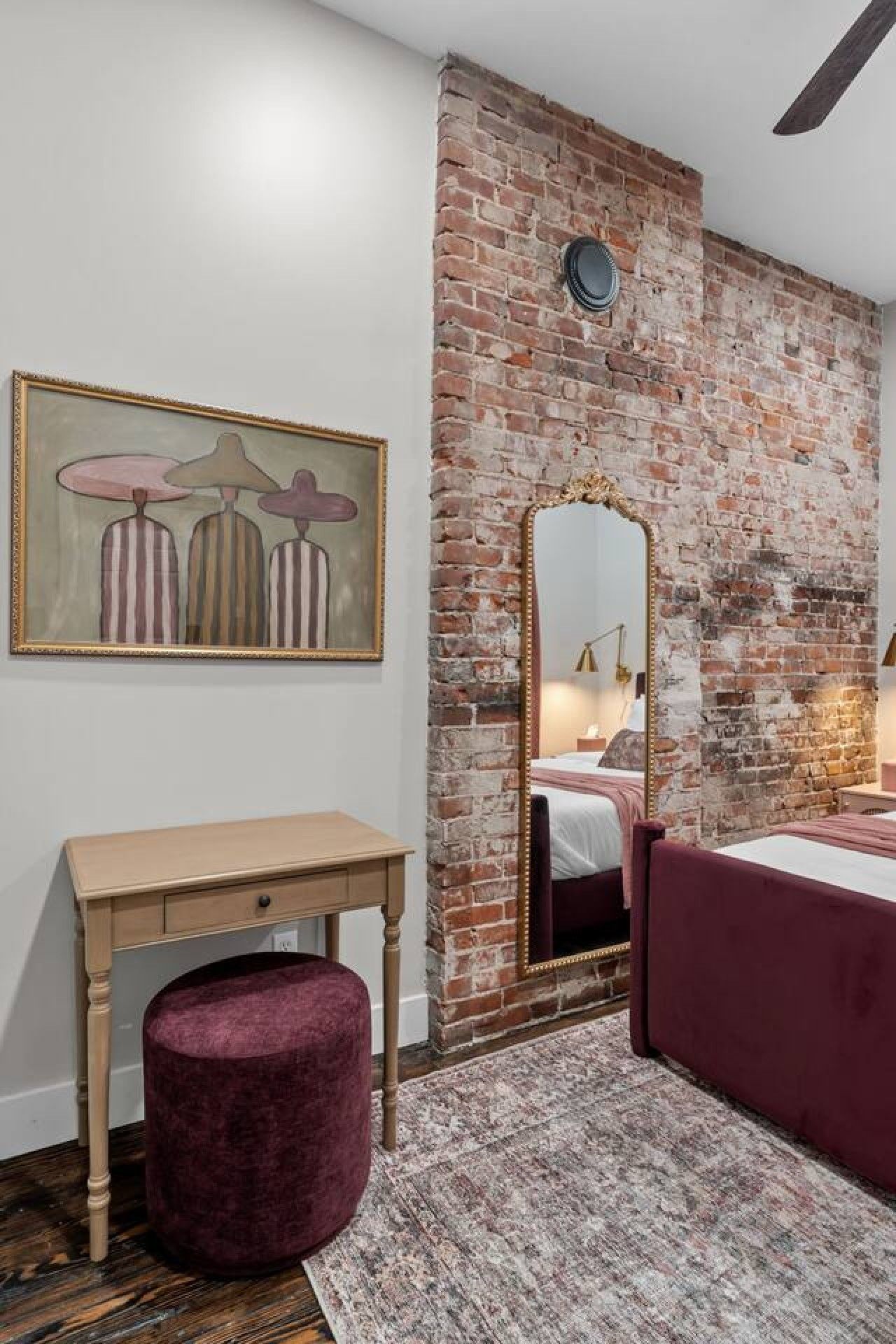 A bedroom with a brick wall, a bed, a nightstand, a mirror, and a painting.