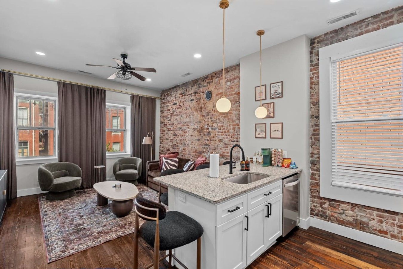 A modern kitchen with a brick accent wall, a dining area with a round table and chairs, and a large window with blinds.