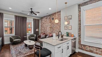 A modern kitchen with a brick accent wall, a dining area with a round table and chairs, and a large window with blinds.