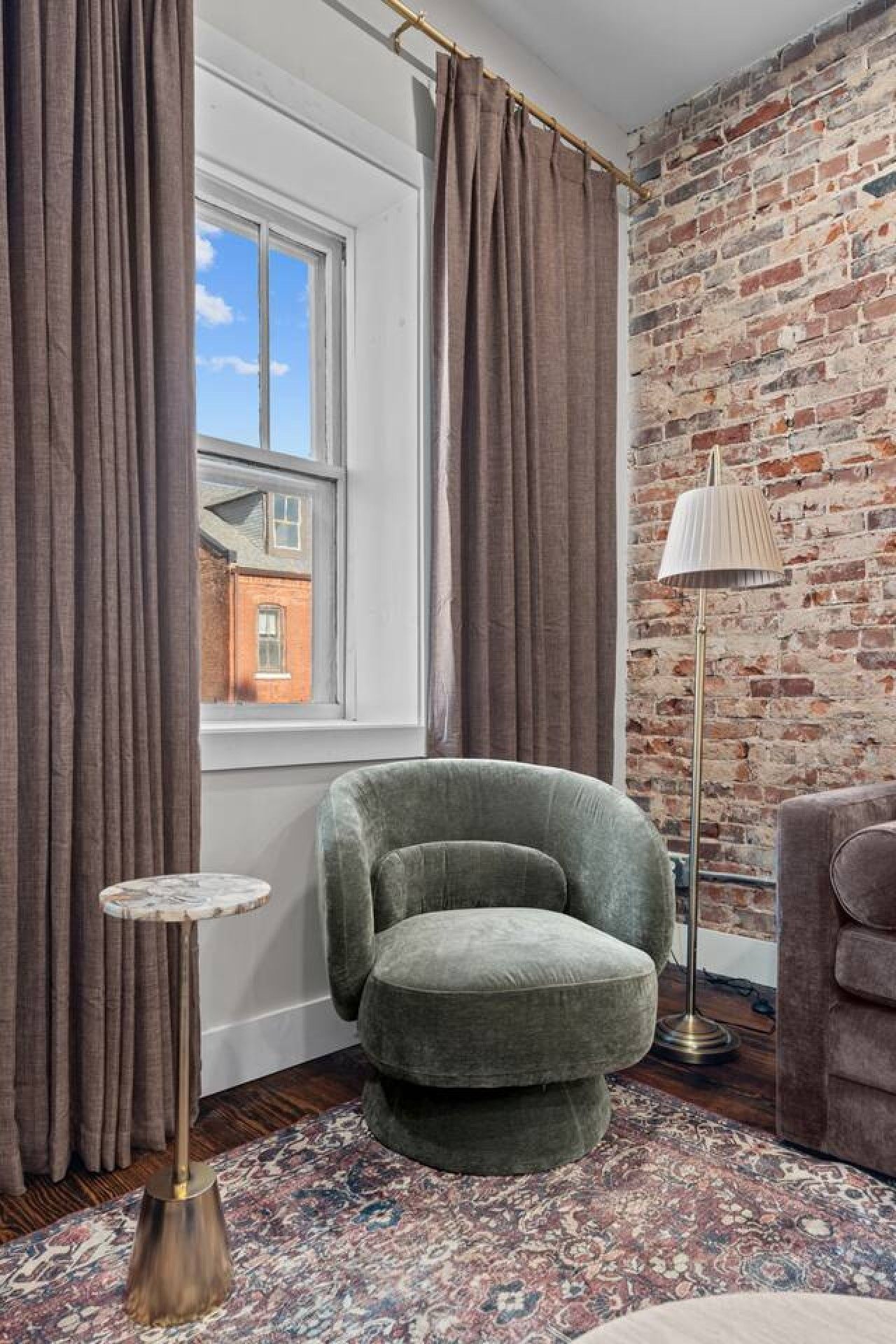 Una habitación con pared de ladrillo, una ventana con cortinas, un sillón verde, una lámpara de pie y una alfombra estampada.