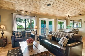 Ein gemütliches Wohnzimmer mit Holzboden, zwei Sofas mit gemusterten Kissen, einem Couchtisch mit einer Vase und großen Fenstern mit Vorhängen.