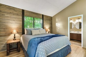 Ein Schlafzimmer mit einem Bett, zwei Nachttischen, einem Fenster mit Jalousien, einem Spiegel und einem Badezimmer mit Waschbecken und Spiegel.