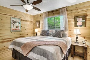 A wooden-paneled bedroom with a bed, two bedside lamps, a ceiling fan, and a poster on the wall. - Smoky Mtn Escape | Hot Tub • Fire Pit • Game Room (Gatlinburg)