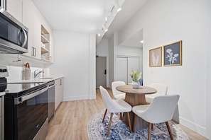 Una cocina moderna con electrodomésticos integrados, una zona de comedor con mesa y sillas redondas, y un pasillo que conduce a un dormitorio.