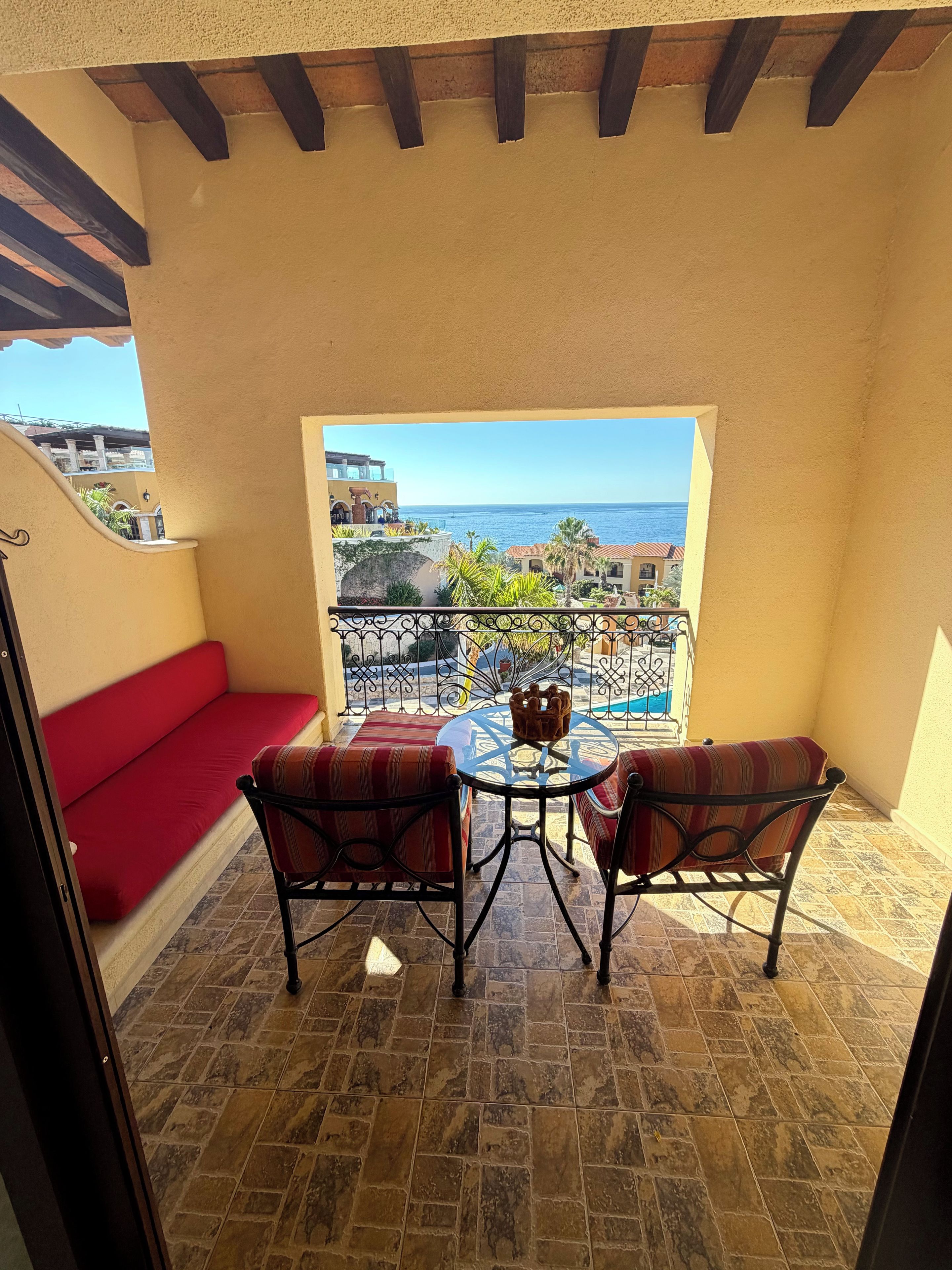 Een balkon met een rode, beklede bank, twee metalen stoelen, een kleine ronde tafel en uitzicht op zee.