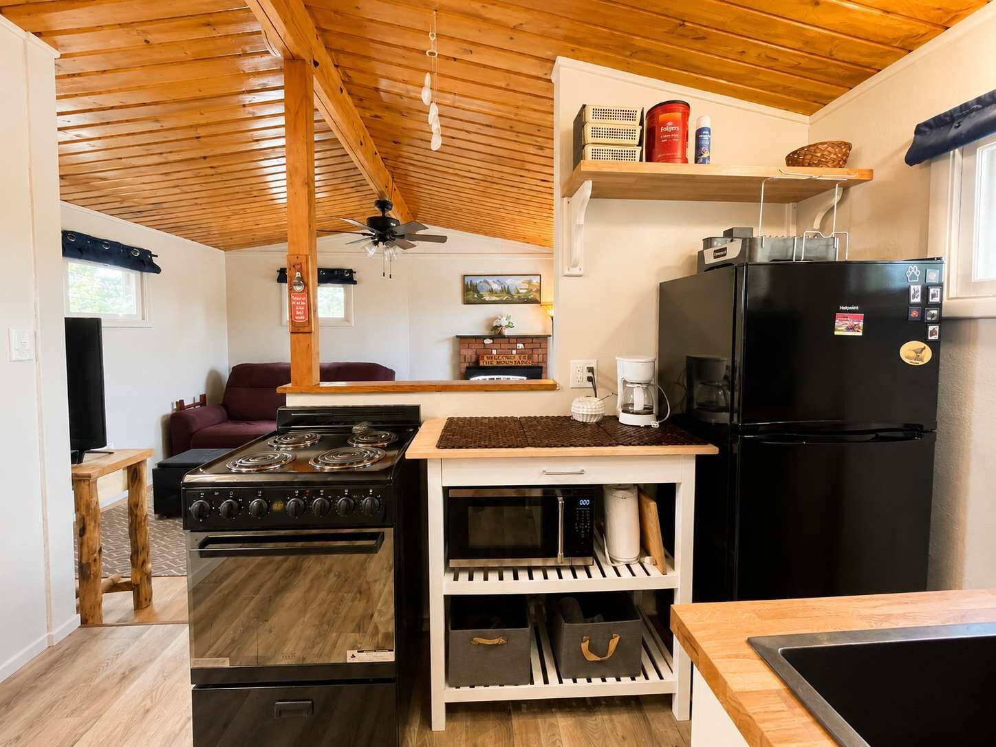 A compact kitchen with a stove, microwave, and refrigerator, wooden ceiling, and a small seating area with a couch.