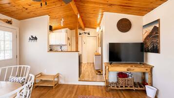 A cozy interior with a wooden ceiling, a dining area with a white chair, a TV on a wooden stand, and a kitchen in the background.