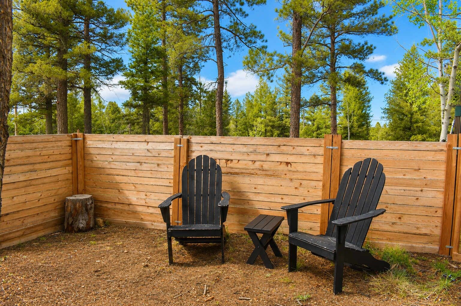 A wooden deck with two Adirondack chairs and a small table, surrounded by a wooden fence and tall trees.