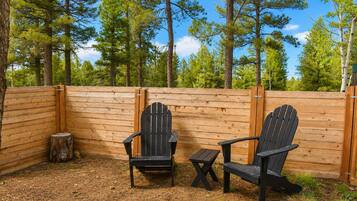 A wooden deck with two Adirondack chairs and a small table, surrounded by a wooden fence and tall trees.