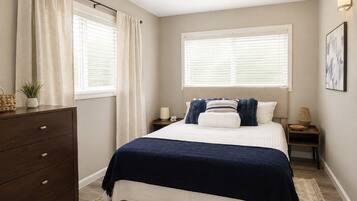 A bedroom with a bed, a dresser, a nightstand, a lamp, and a window with curtains.