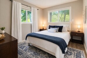 A bedroom with a large bed, two bedside tables with lamps, a window with blinds, a patterned rug, and a nightstand with a basket.