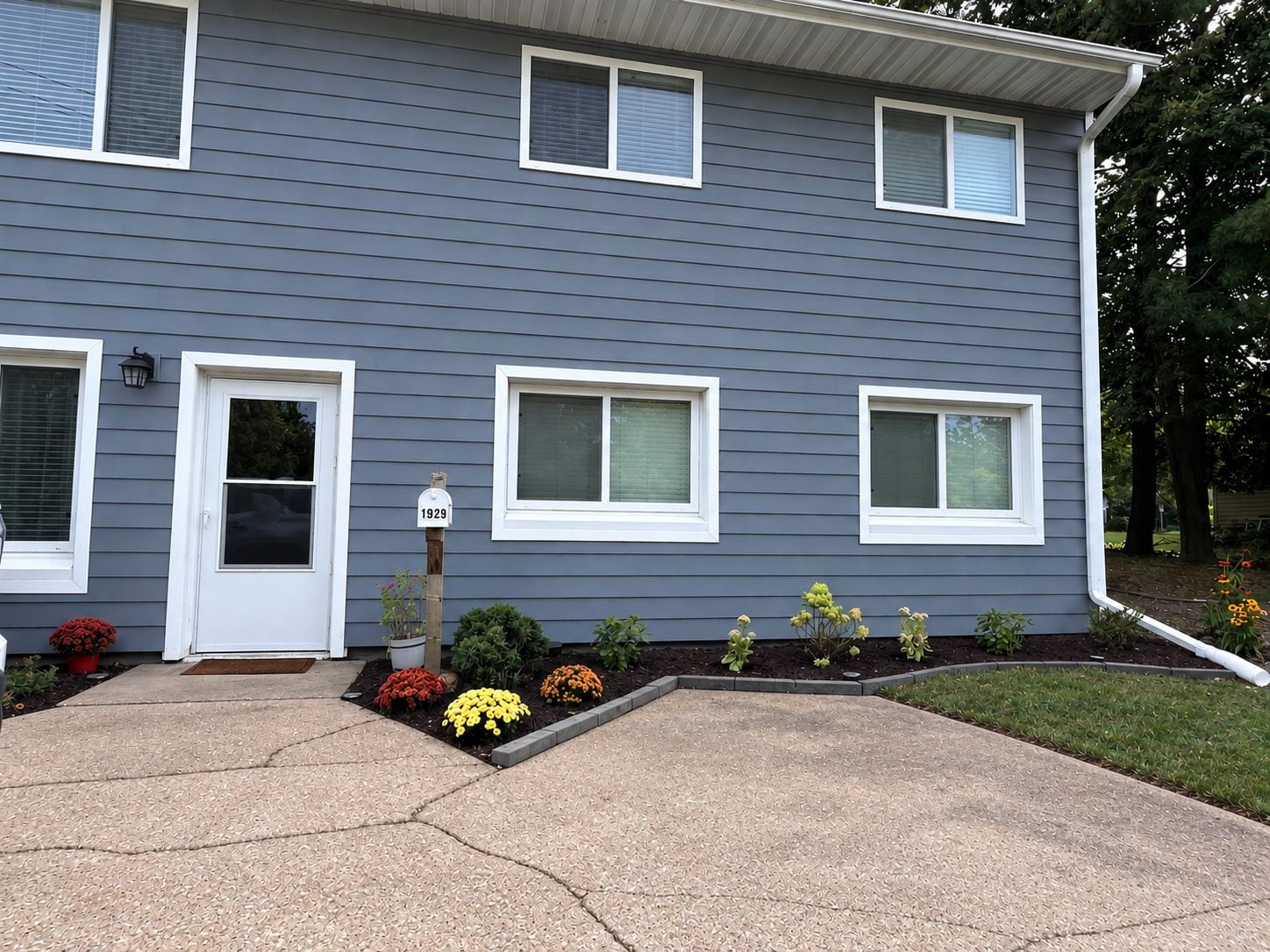 A two-story residential building with a gray exterior, white trim, and a front door. There are three windows on the ground floor and two on the upper floor. A mailbox with the number 1929 is visible. The front yard has a variety of flowering plants and shrubs.