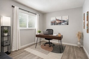 A modern office space with a desk, chair, and laptop, a window with curtains, a plant, and artwork on the wall.