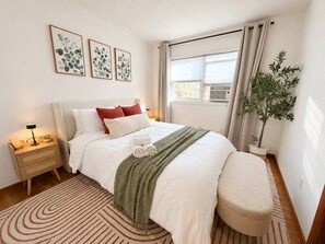 A hotel room with a bed, bedside table, lamp, window, curtains, and a potted plant. - A Rare, Spacious Retreat - Downtown JC, Free Parking, Close to PATH (Jersey City)