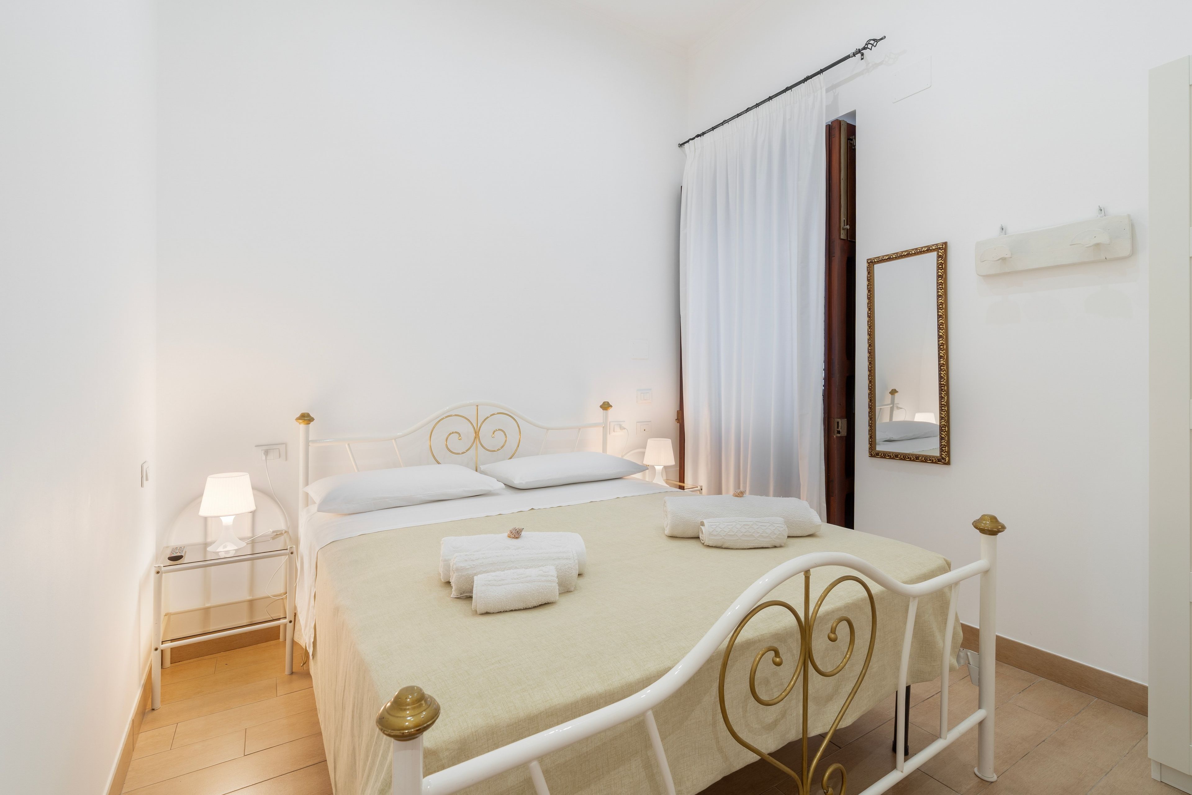 A neatly made bed with white linens and a beige blanket, a bedside table with a lamp, and a mirror on the wall.