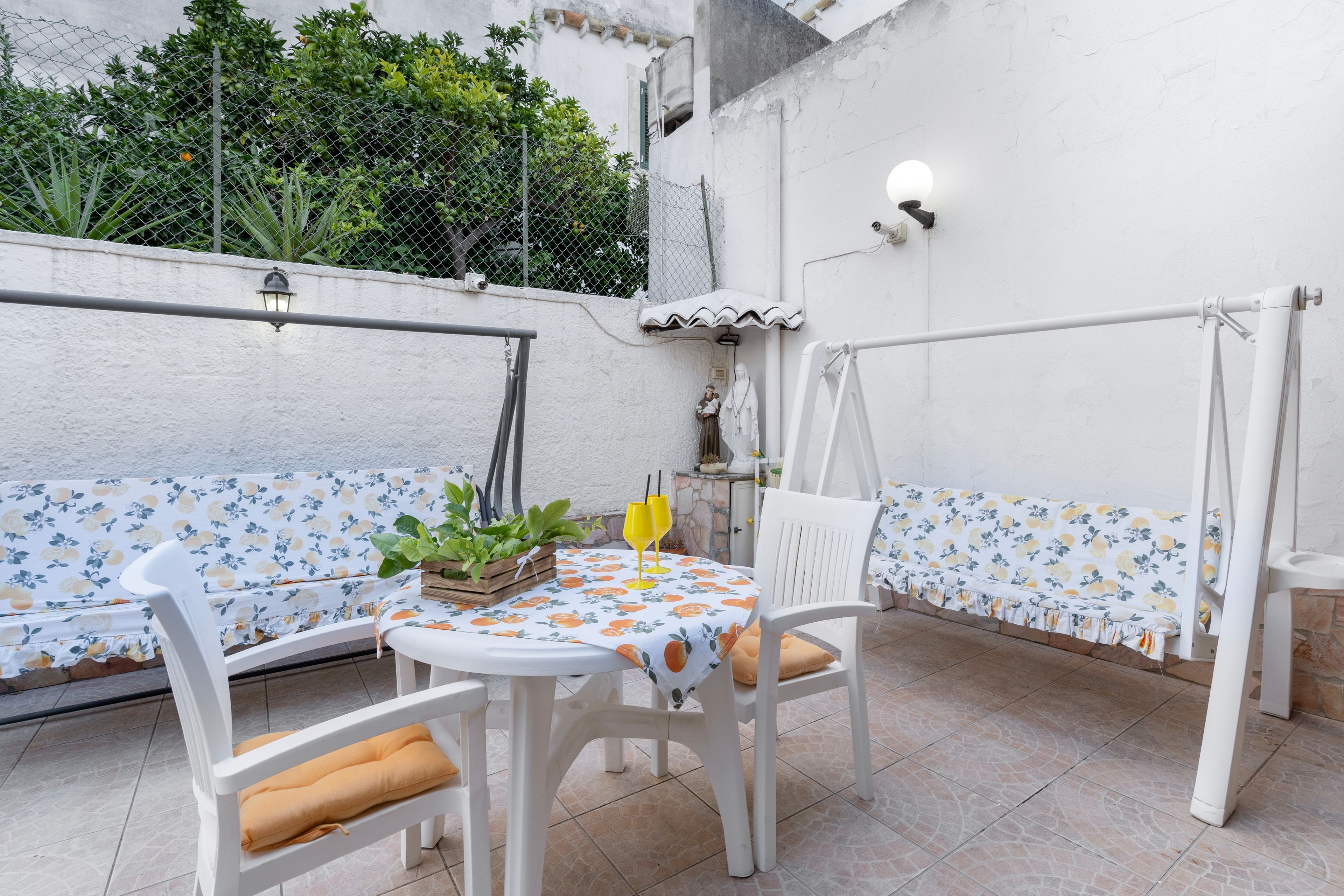 An outdoor seating area with white chairs and a table, surrounded by a white wall and a metal fence.
