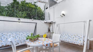 An outdoor seating area with white chairs and a table, surrounded by a white wall and a metal fence.