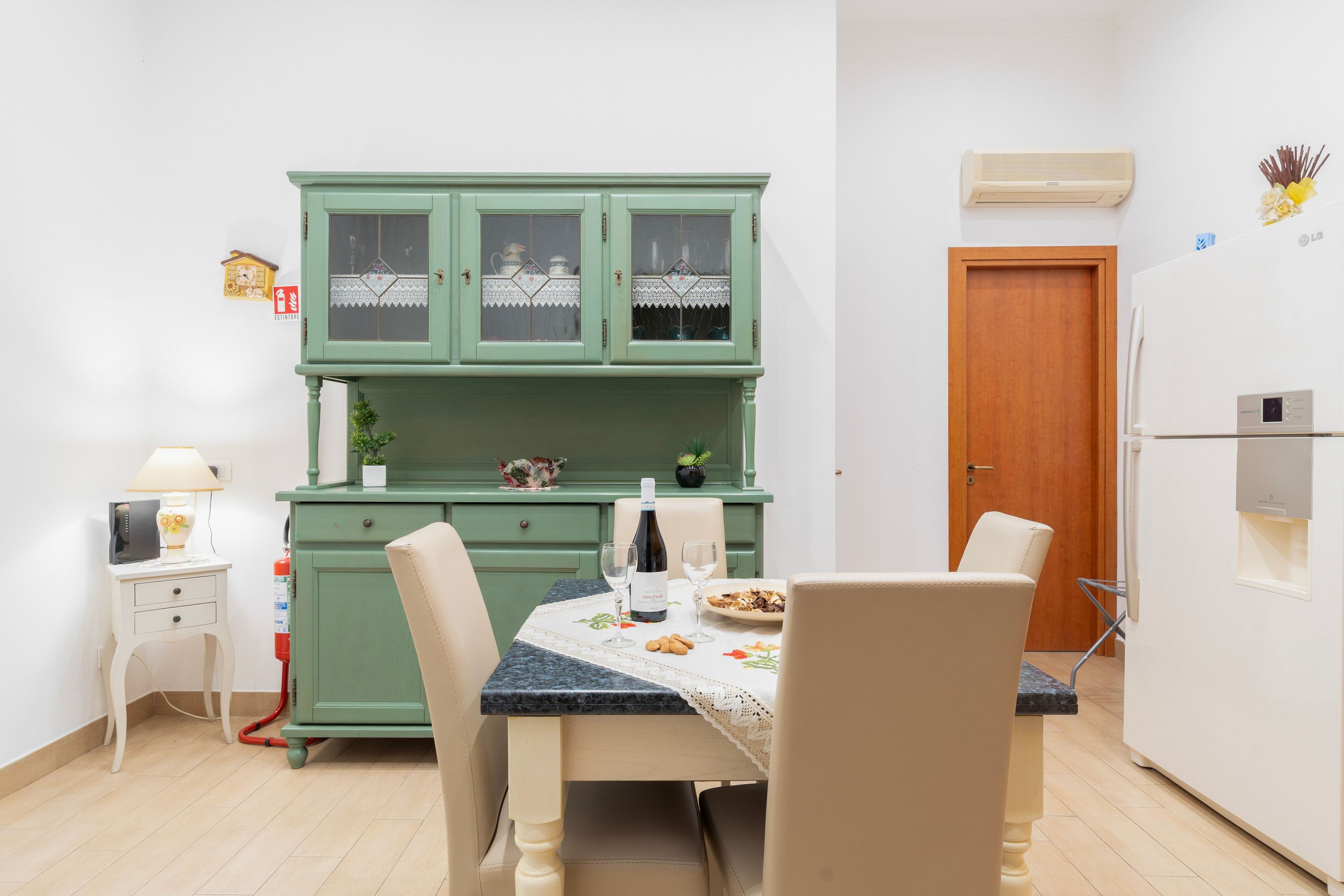 A compact kitchen with a green cabinet, a dining table with chairs, and a small side table with a lamp.