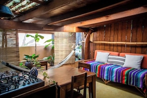 A cozy room with a wooden table, a sofa with colorful cushions, and a large window with plants. - La Siesta Troncones    Beautiful Cozy Palapita (Troncones)