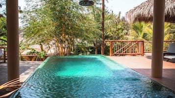 A pool area with a thatched roof structure, surrounded by greenery and palm trees.