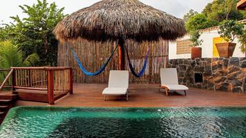 A pool area with a thatched-roof structure, two white lounge chairs, and a wooden deck with a railing.