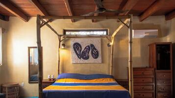A bedroom with a four-poster bed, a ceiling fan, a window with natural light, a wall-mounted artwork, and wooden furniture.