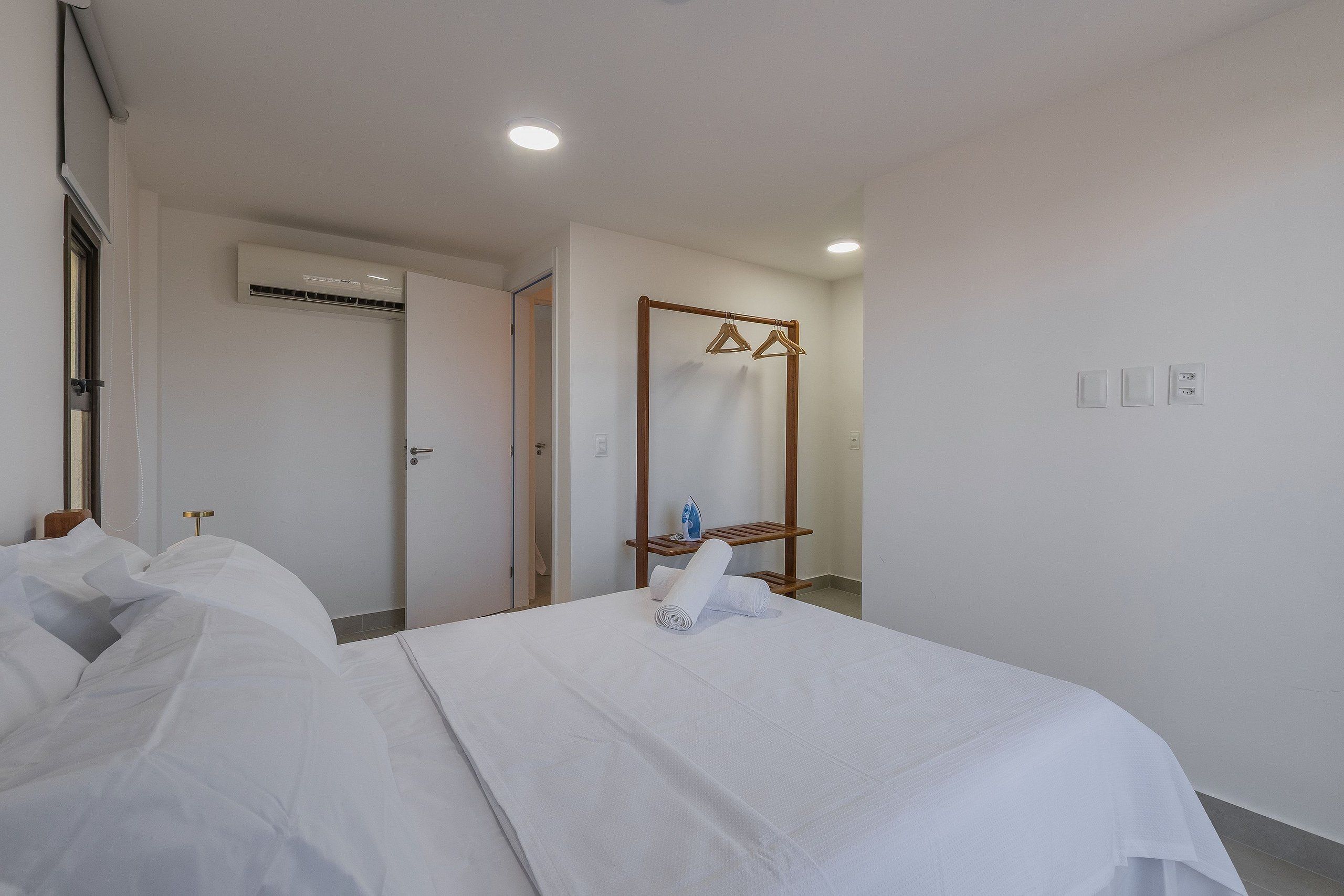 Habitación de hotel con cama, perchero de madera, unidad de aire acondicionado y puerta que da a otra habitación.