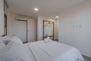 Habitación de hotel con cama, perchero de madera, unidad de aire acondicionado y puerta que da a otra habitación.
