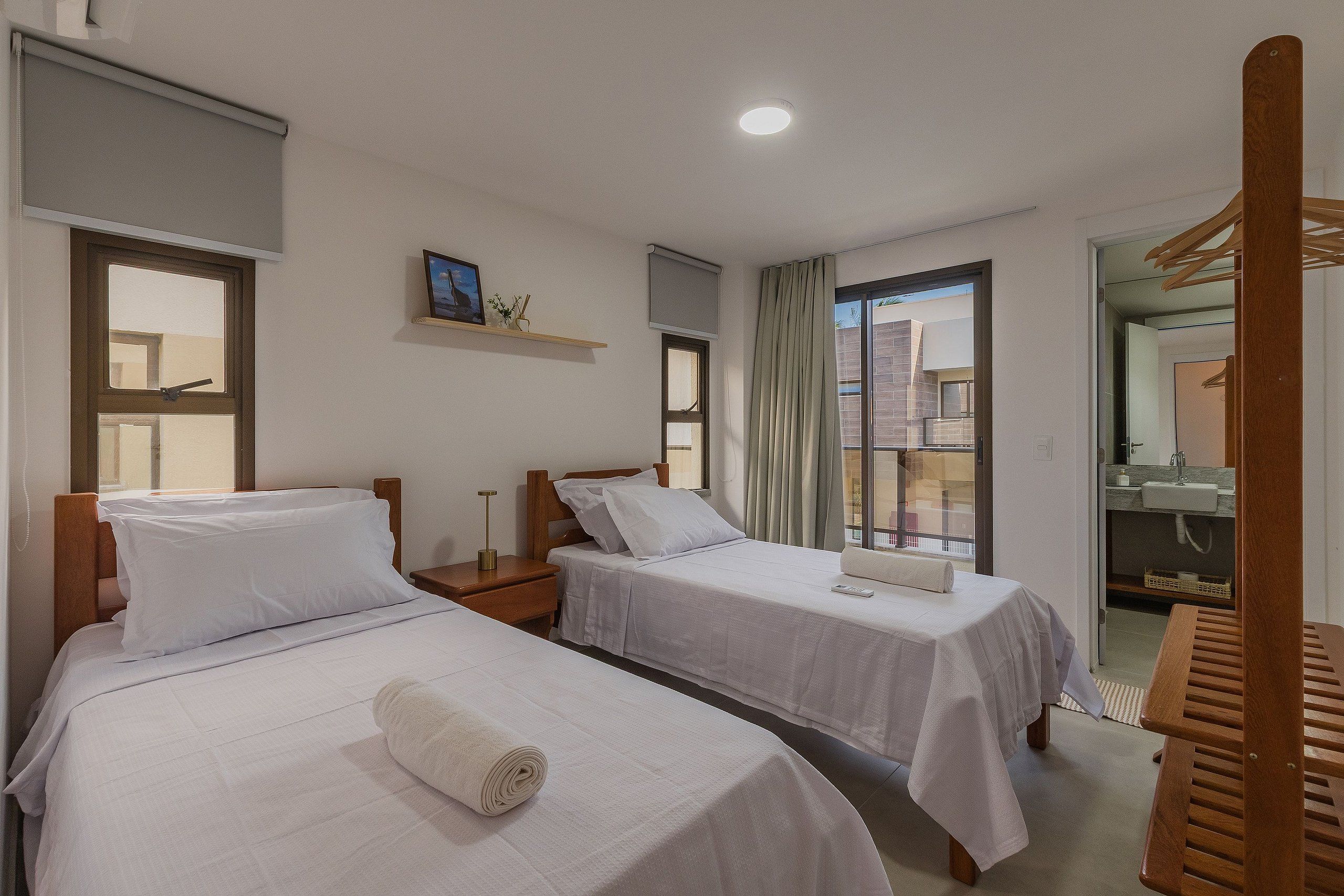 A hotel room with two beds, a small bedside table with a lamp, a window with curtains, a shelf with a picture, and a view of the outside.