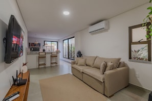 A living room with a flat-screen TV, a sofa, a coffee table, and a kitchen area with a refrigerator and a microwave. - Villa Vento #c03 - House in Cumbuco by Carpediem (Caucaia)