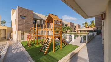 Un parque infantil con una estructura de madera para escalar y un tobogán, rodeado de césped cuidado y una zona de piscina.