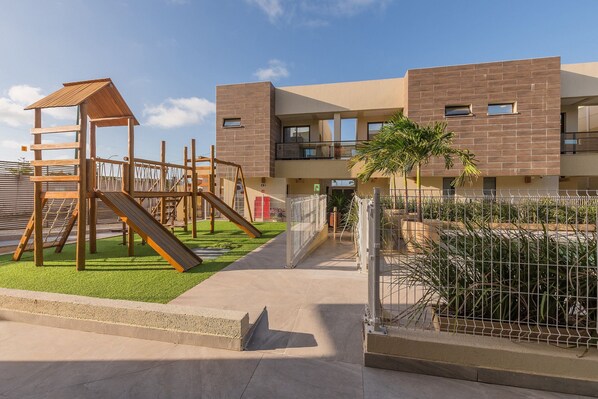 Un parque infantil con un tobogán de madera y estructuras para escalar, junto a un edificio moderno con balcones.