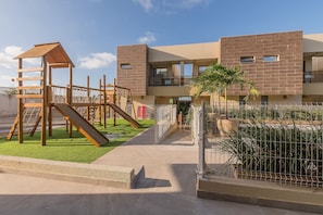 A playground with a wooden slide and climbing structures, adjacent to a modern building with balconies.