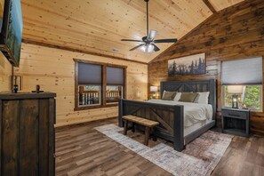 Une chambre à coucher avec un lit, un ventilateur de plafond, une fenêtre avec des stores, une table de chevet et un plancher en bois.