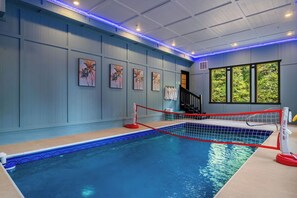 An indoor swimming pool with a red net, surrounded by walls with artwork, a staircase, and a window with a view. - New Luxury Lodge • Private Pool • Hot Tub • Game Room • Sleeps 18 (Sevierville)