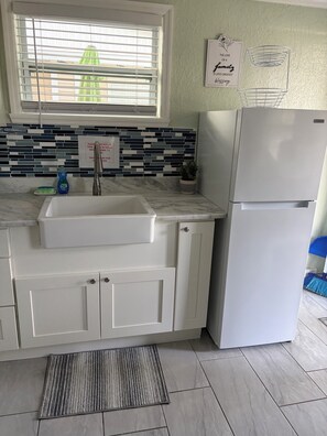 A kitchen with a white sink, a refrigerator, a window with blinds, a tiled backsplash, and a sign on the wall.
