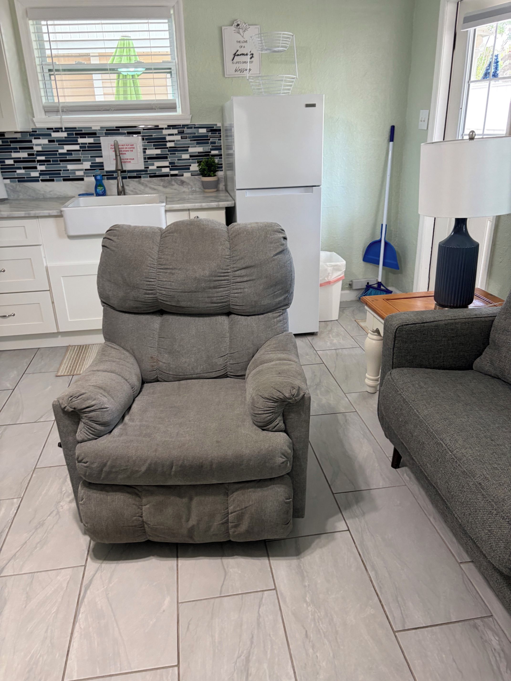 A living room with a grey sofa, a grey armchair, a white refrigerator, and a window with blinds.