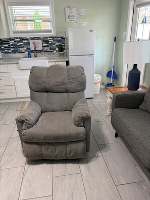 A living room with a grey sofa, a grey armchair, a white refrigerator, and a window with blinds.