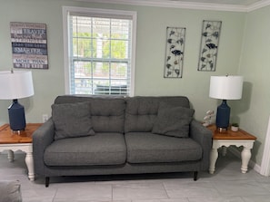 A living room with a grey sofa, two side tables with lamps, a window with blinds, and wall art.