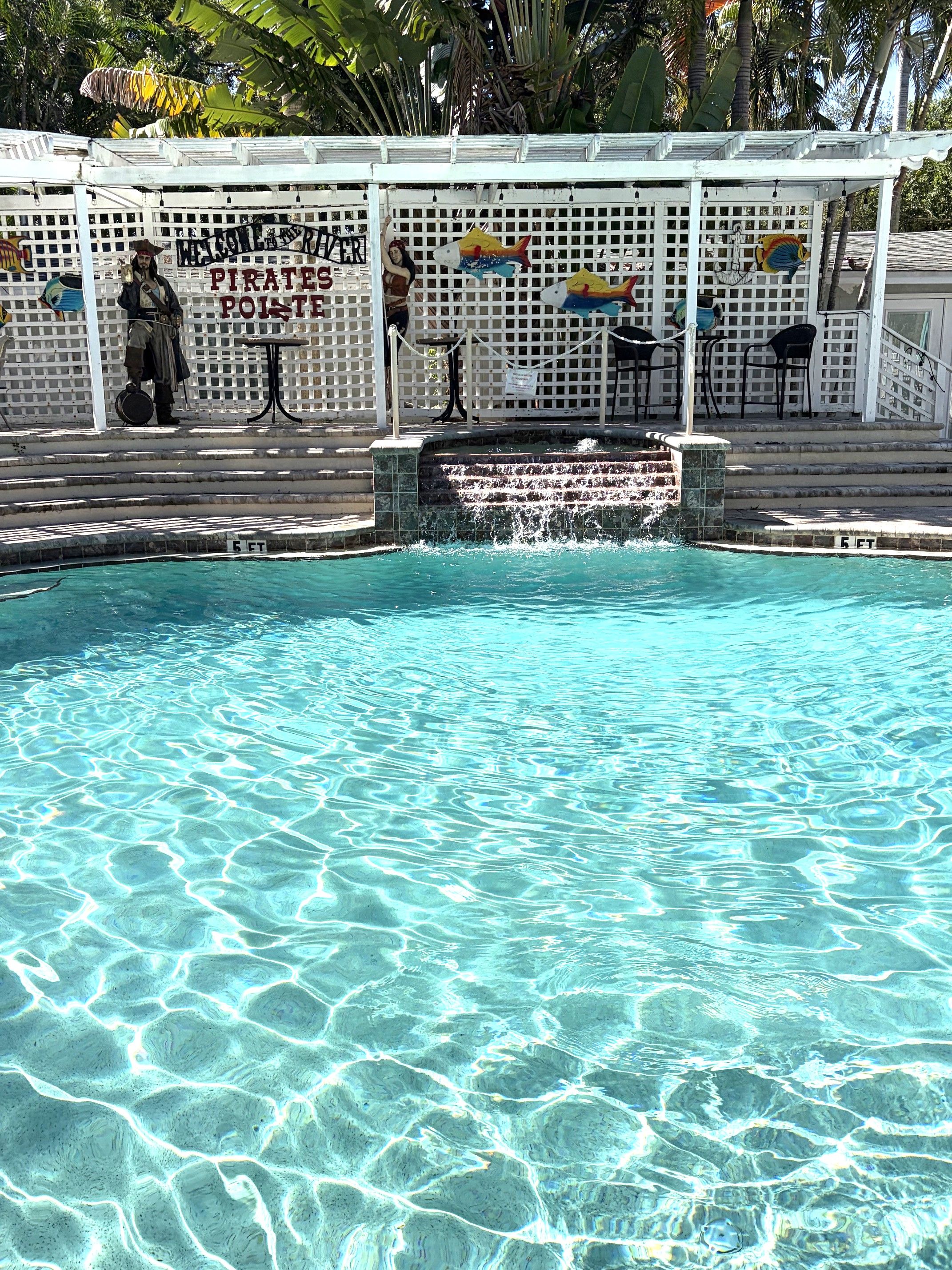 Una piscina cristalina con cascada, rodeada por una cerca blanca decorada con peces y una estatua de pirata.