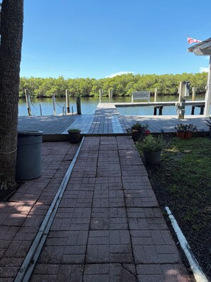 A wooden dock leading to a calm body of water, surrounded by greenery and a clear sky.