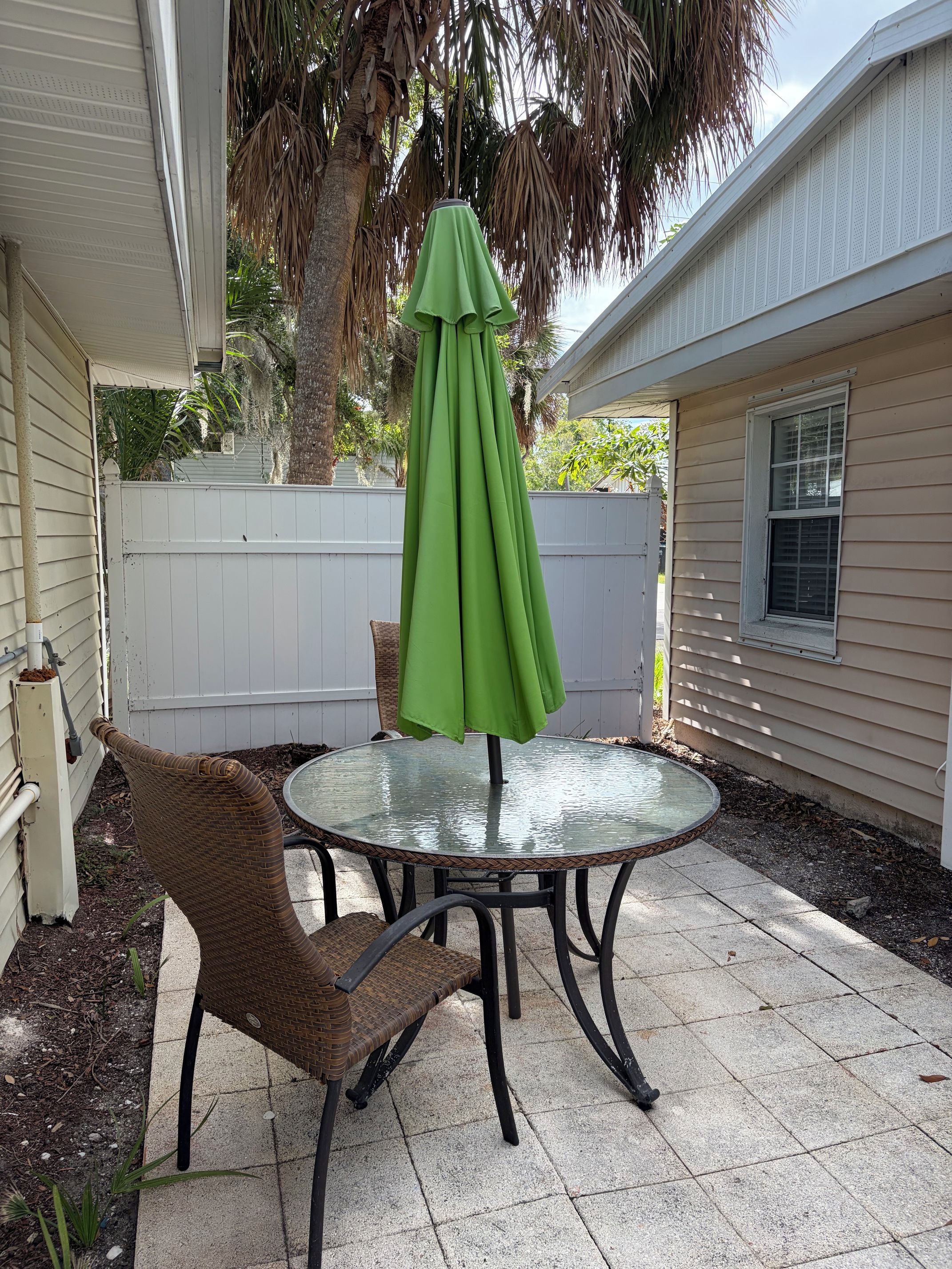 A patio with a round glass table and chairs, a green umbrella, and a white fence.