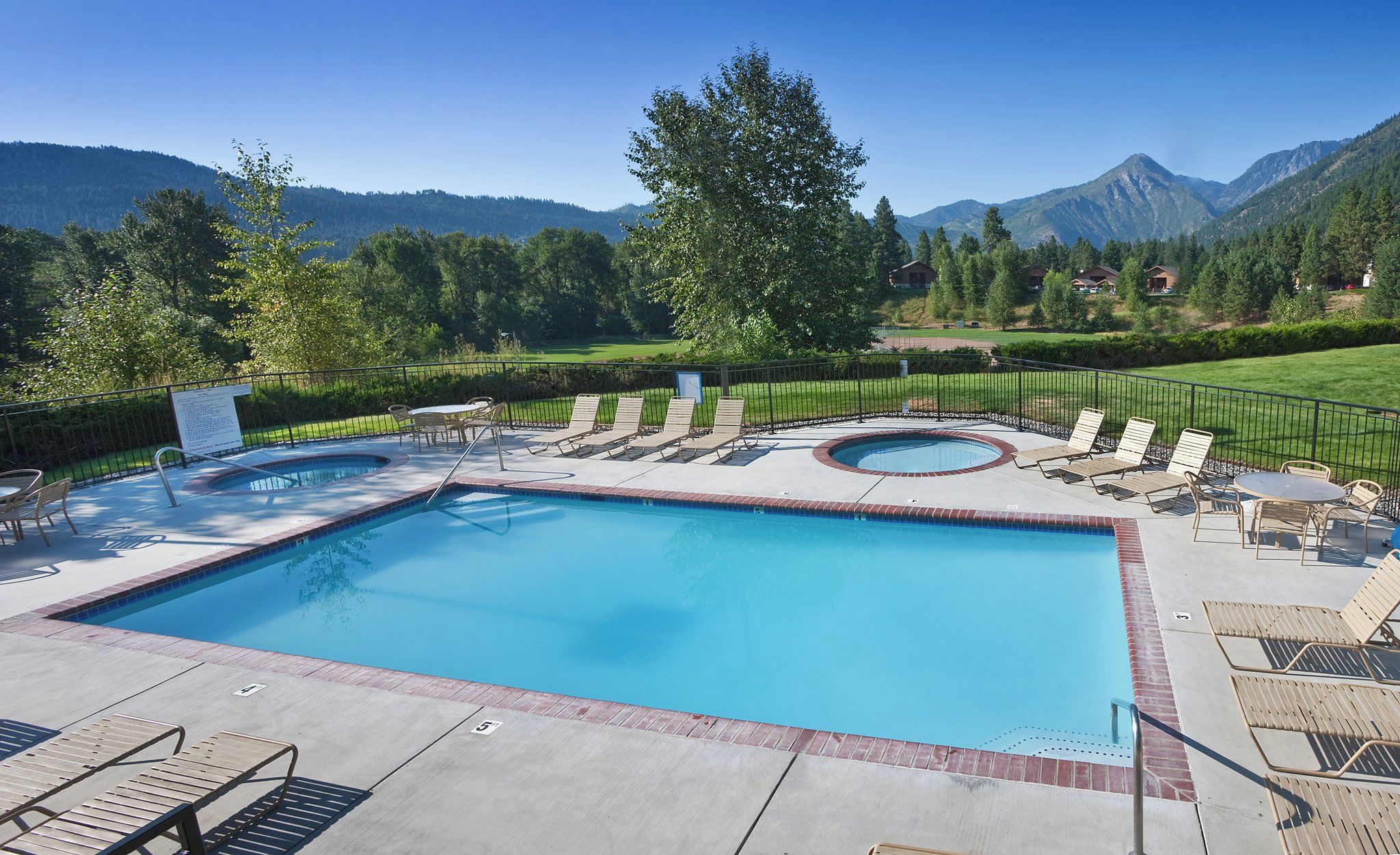 Ein Swimmingpool mit Liegestühlen und einer Whirlpool, umgeben von einem Zaun und Bäumen, mit Bergen im Hintergrund.