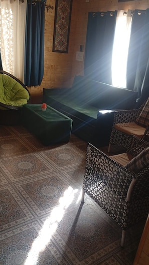Una habitación con una cama, una silla tapizada de verde, un sillón estampado y una planta pequeña.