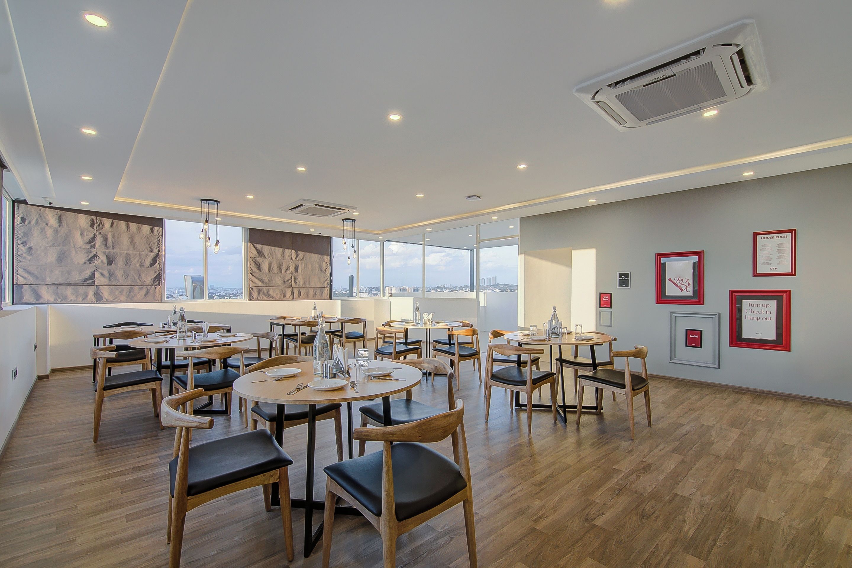 A spacious dining area with multiple tables and chairs, large windows offering a city view, and a wall with framed artwork.