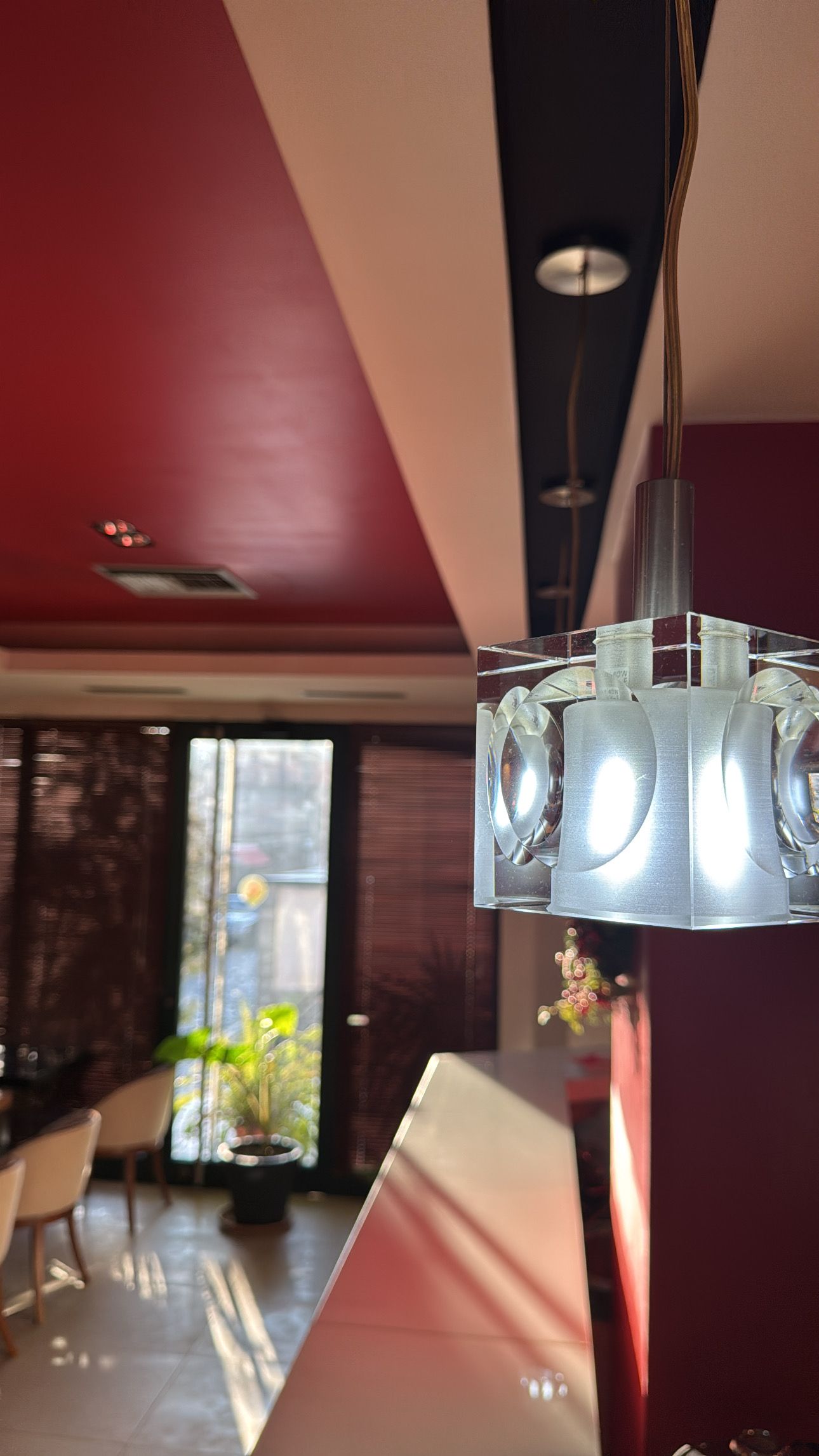 Un intérieur moderne avec un luminaire suspendu en verre transparent, un mur rouge et une vue sur un coin repas avec des chaises et une table.