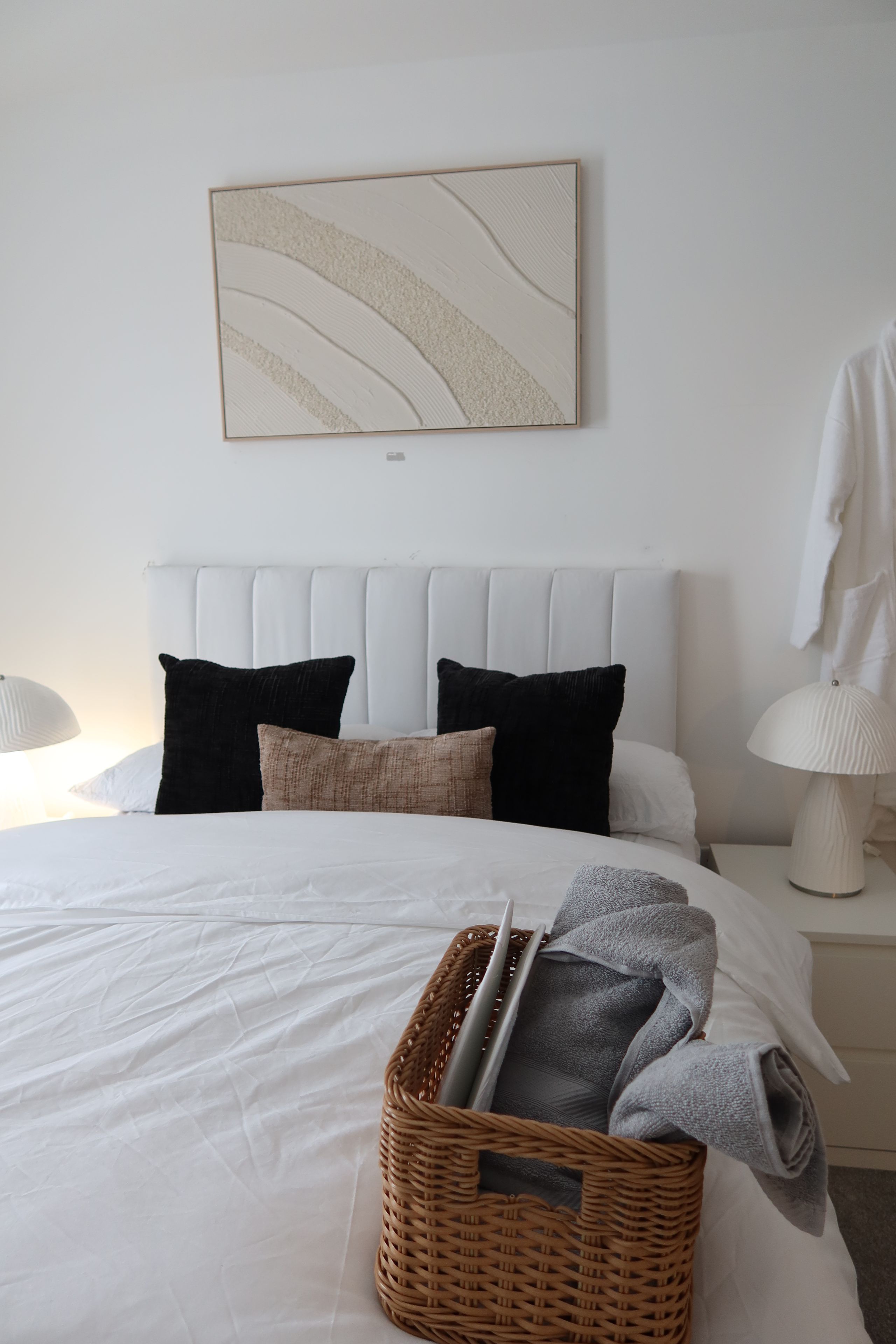 Un letto rifatto con un comofort bianco, una testiera con numerosi cuscini, un'opera d'arte astratta appesa al muro, un cesto di vimini con asciugamani piegati e un laptop, e una lampada da comodino.