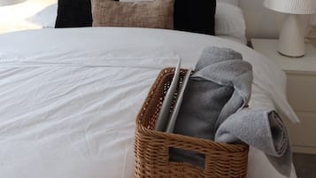 Un letto rifatto con un comofort bianco, una testiera con numerosi cuscini, un'opera d'arte astratta appesa al muro, un cesto di vimini con asciugamani piegati e un laptop, e una lampada da comodino.