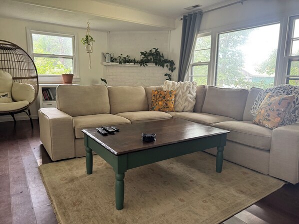 Ein Wohnzimmer mit einer Eckcouch, einem Couchtisch, einer hängenden Pflanze und einem Fenster mit Vorhängen.