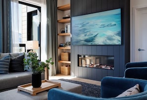 A modern living room with a sofa, a blue armchair, a fireplace, and a large TV mounted on the wall. - 2 Turns by Revelstoke Vacations (Revelstoke)